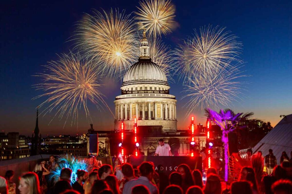 A rooftop party with people dancing, a DJ performing, and colourful fireworks lighting up the night sky above St Paul’s Cathedral in London. The scene is festive with bright lights and palm decorations.